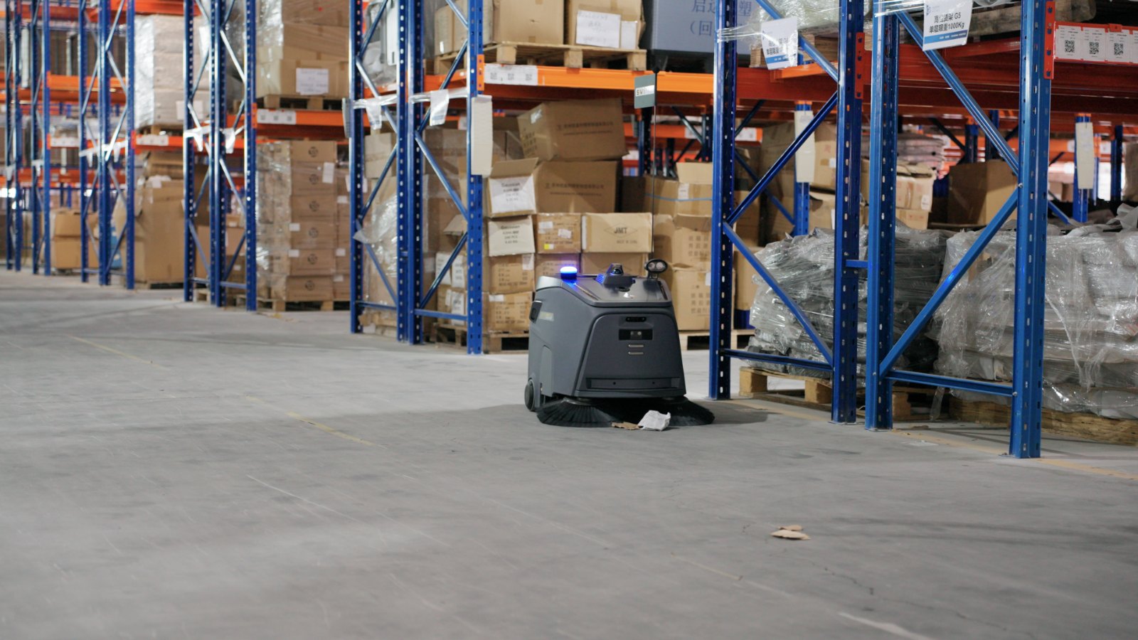 Gausium Beetle sweeping robot cleaning the floor of a warehouse aisle between pallet racking