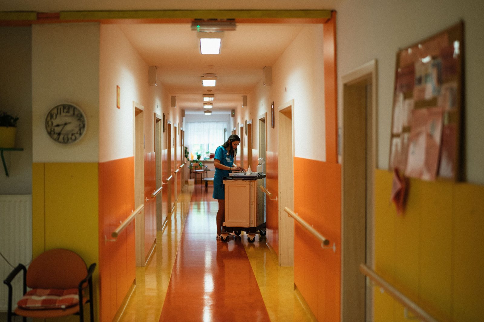 A care worker attends a medication cart in a brightly lit aged care facility corridor